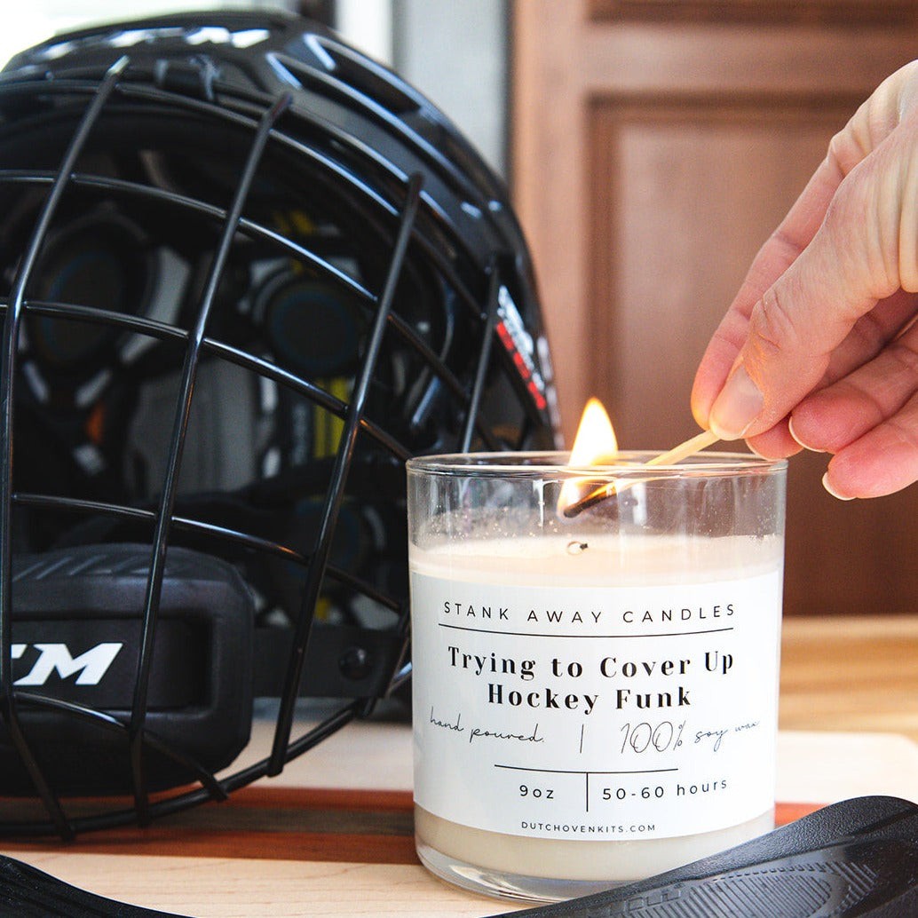 A woman's hand lights a white Stank Away soy wax candle cinnamon vanilla scent. Trying to cover up hockey funk. The candle is on a table with a hockey helmet in the background.
