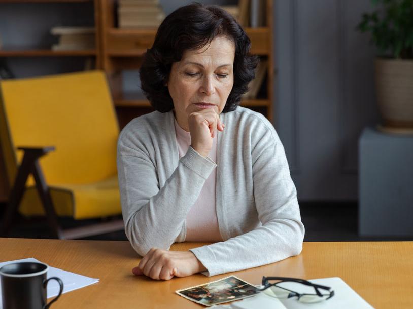 Middle-aged woman sitting at a table, looking thoughtfully at old photographs with a serious expression. Glasses, a mug, and a piece of paper are on the table.