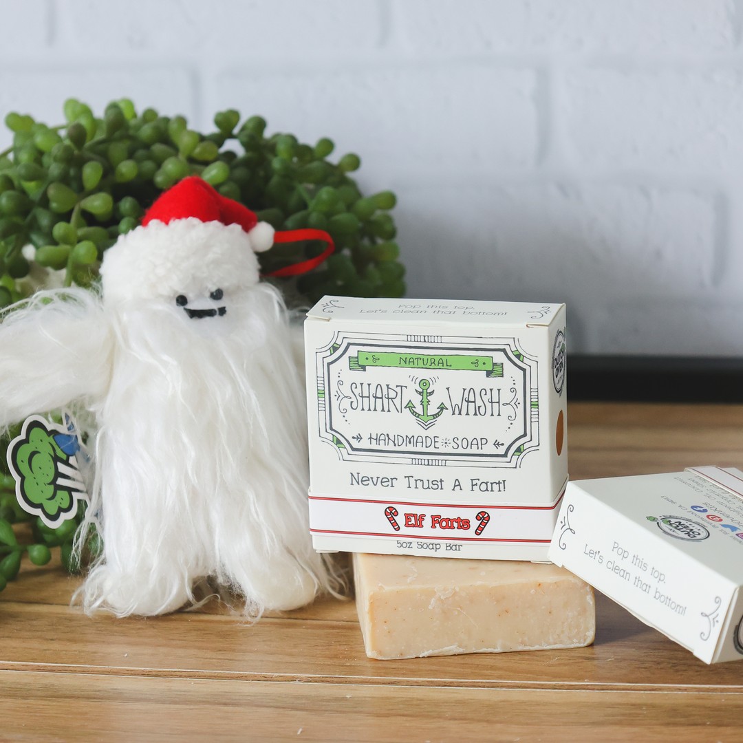 A box of shart wash elf farts scented soap on top of a tan soap bar on a wood table with a christmas yeti. Oatmeal spice scented holiday soap