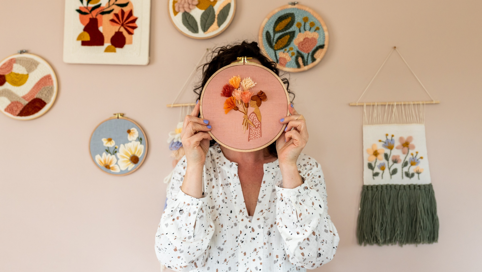 A woman holding a pink embroidery hoop with a stitched bouquet in front of her face, against a wall of hoop art.