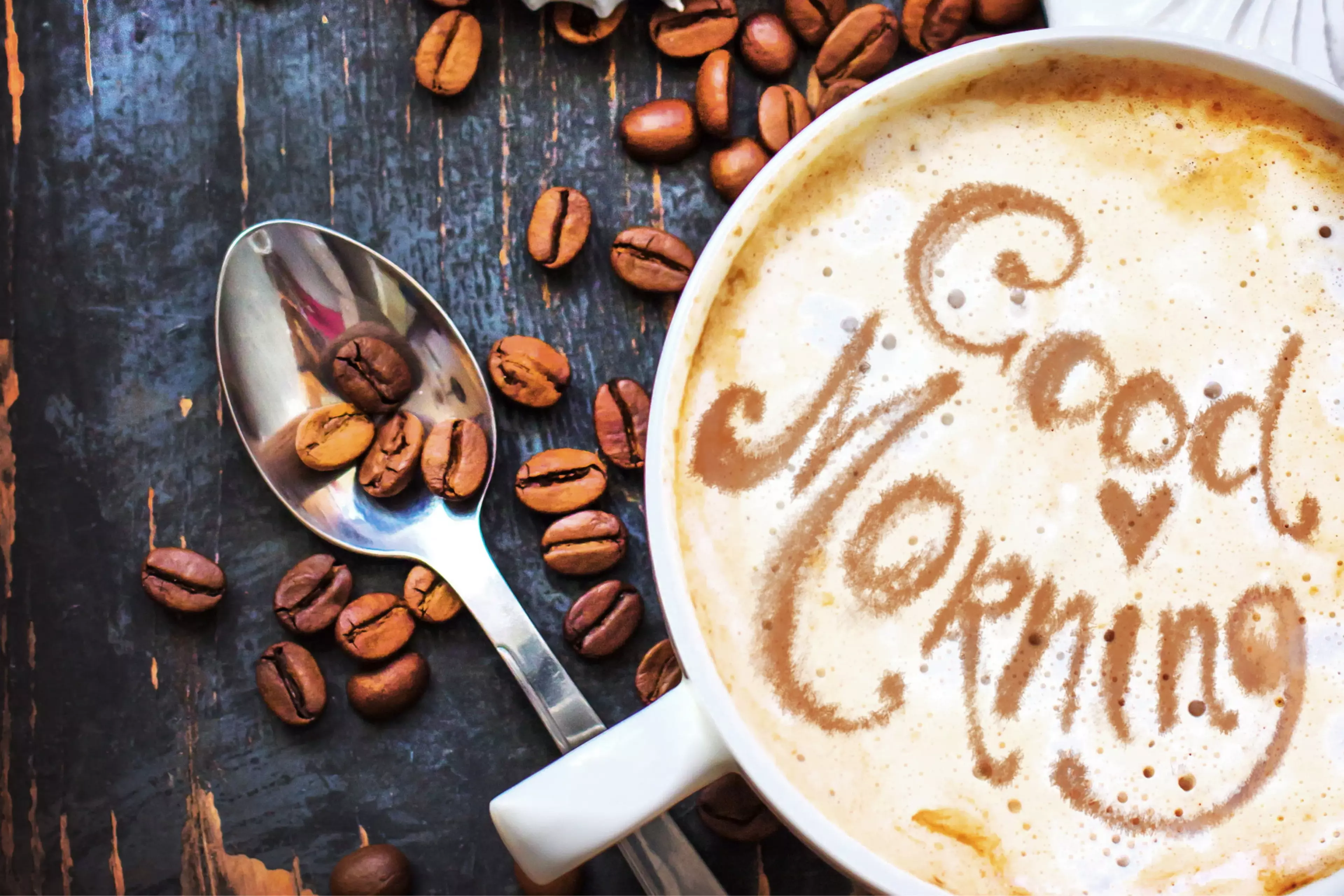 Close-up latte with “Good Morning” written in foam beside scattered coffee beans and a spoon.