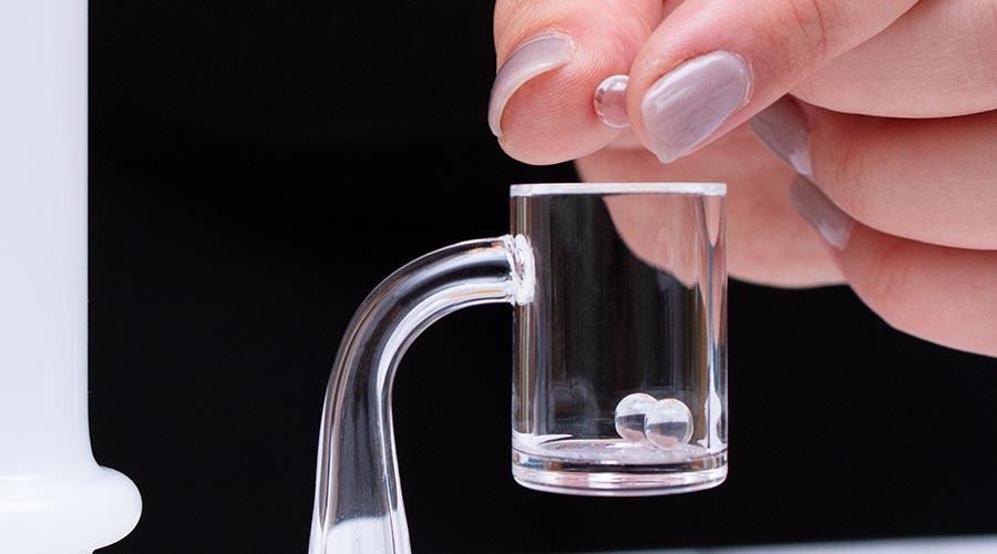 A hand carefully lowering a quartz terp pearl into a clear quartz banger. This image illustrates how to use terp pearls and a carb cap to maximize vapor production and flavor at low temperatures on a Prism modular rig.