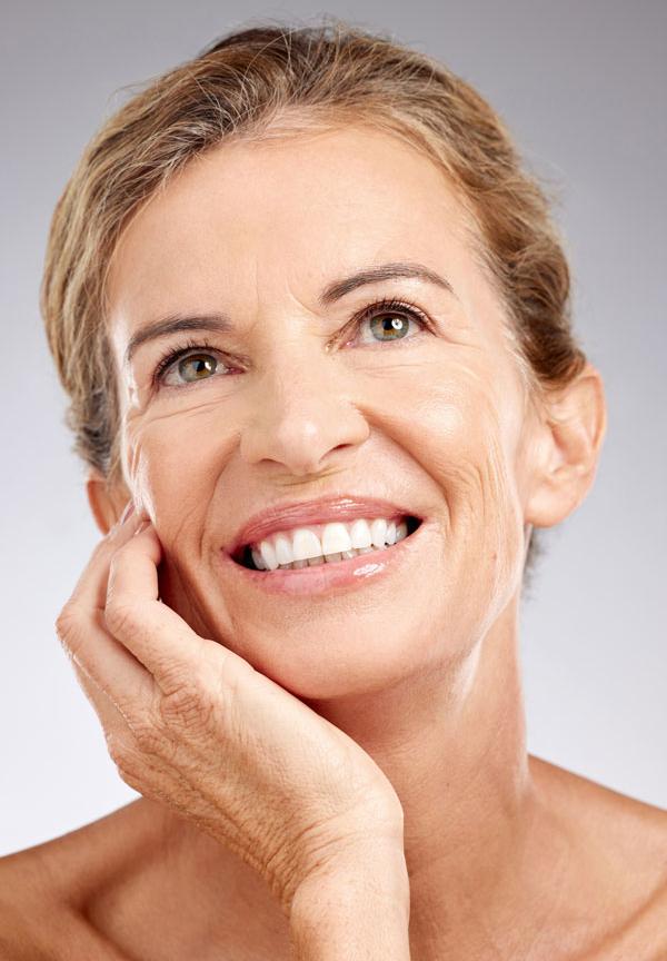 Smiling older woman with glowing skin and her hand resting on her cheek, looking upward with a serene expression.