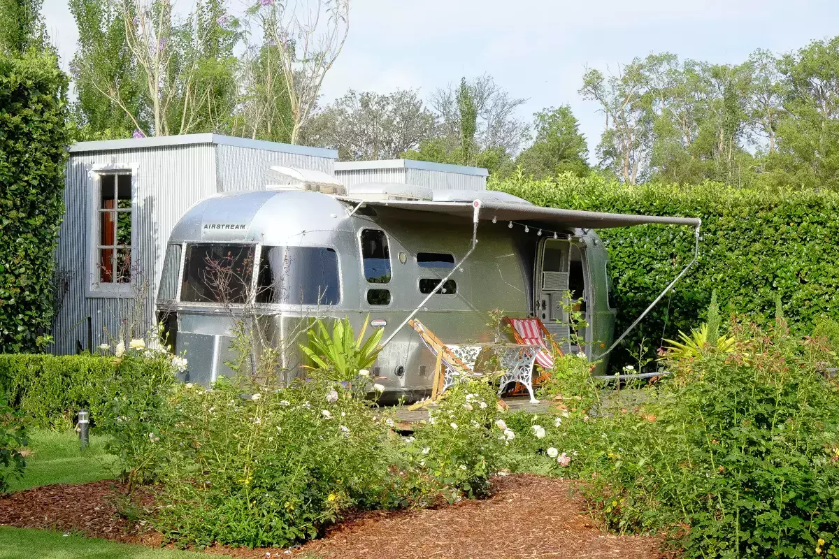 Restored silver Airstream caravan set among lush garden beds, featuring a timber deck with outdoor chairs, retractable awning, and corrugated metal outbuildings surrounded by hedges and trees in a tranquil rural setting.