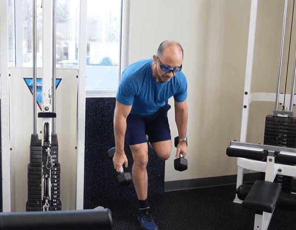 Man In Blue with Shorts Holding Dumbbells in Gym Outside View