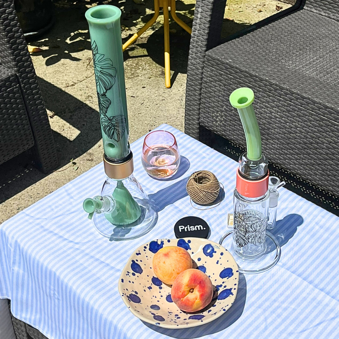 Summer tablescape featuring two Monstera Decal Prism bongs on a striped tablecloth: A Mint Green Percolated Beaker Bong and a Key Lime Green Honeycomb Bong. The scene, complete with peaches and a glass of wine, highlights the decals' fit into a relaxed, biophilic home decor and lifestyle setting.