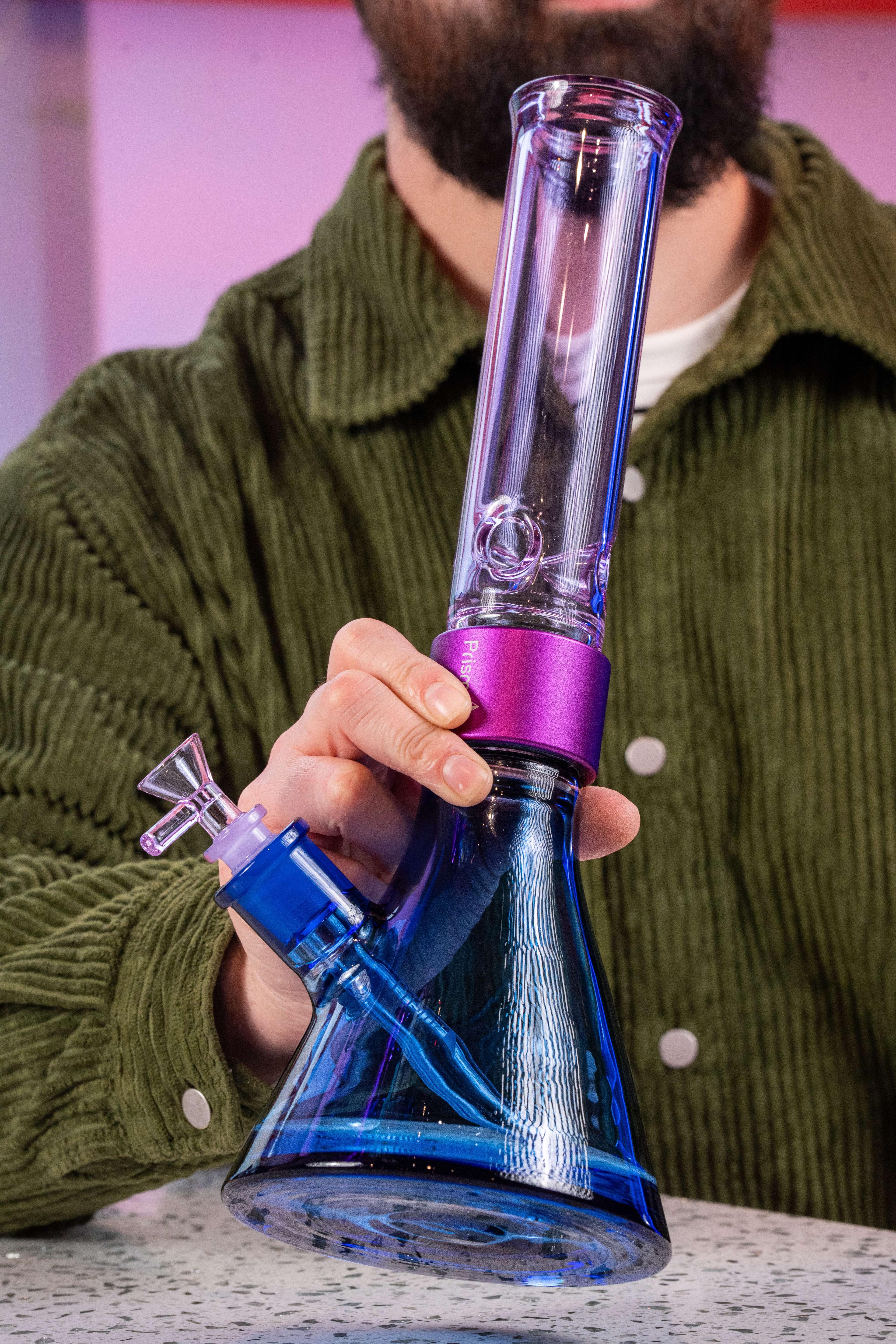 A person holding a blue and purple Prism modular glass beaker pipe from the Duo collection to their mouth, demonstrating the scale and size of the waterpipe. The image highlights the Sapphire Blue base, Grape Jolly Rancher purple mouthpiece, and matching purple Halo Connector in a lifestyle setting.
