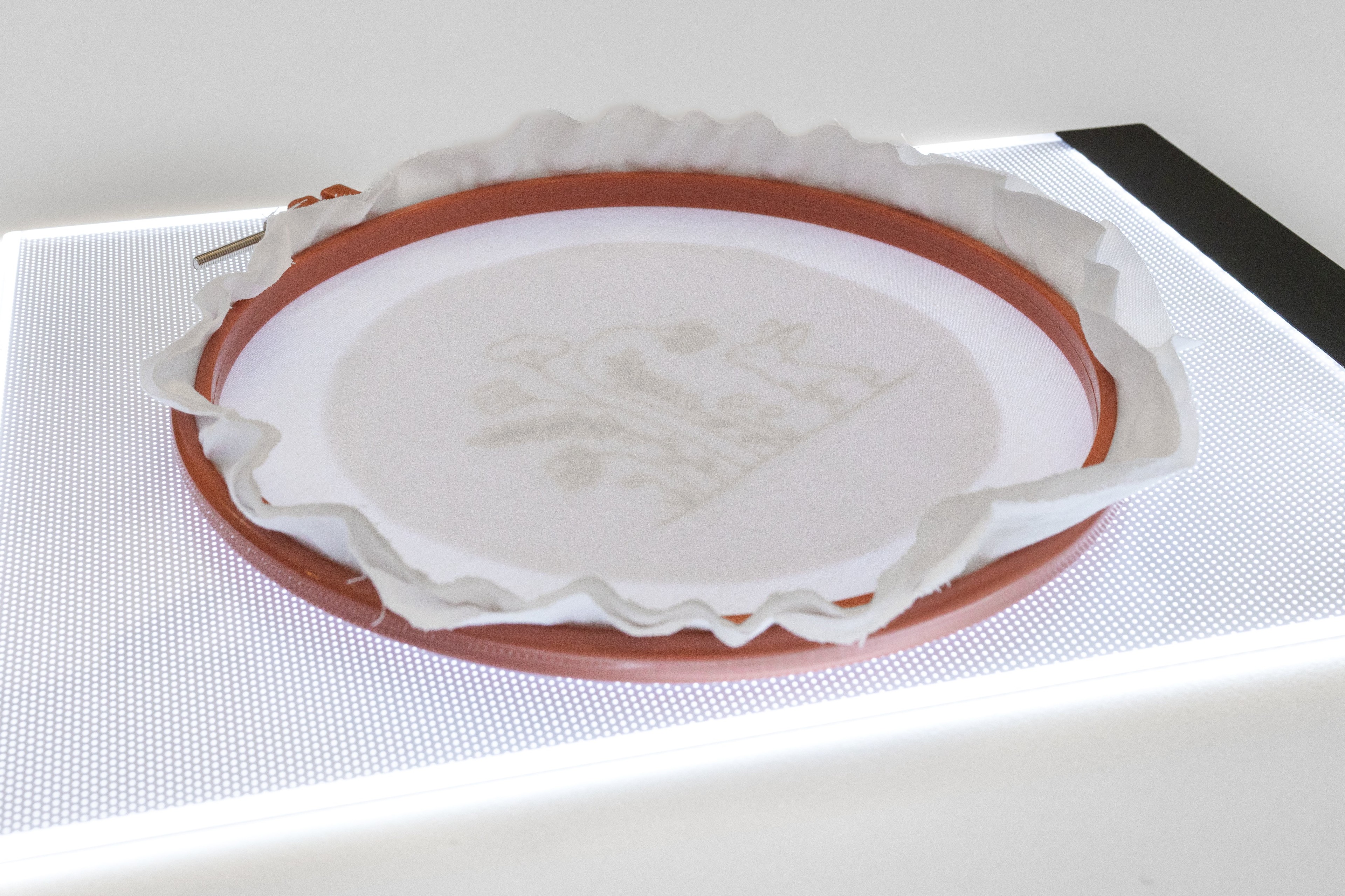 A hoop with a pattern drawn on it and fabric overtop lies on a lightbox.