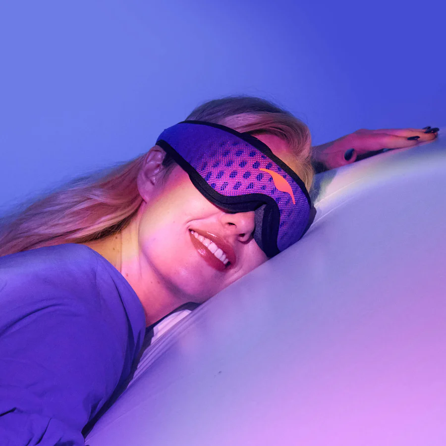 A smiling blonde girl lying down on a white pillow, wearing a blue mesh sleep mask for side sleepers.