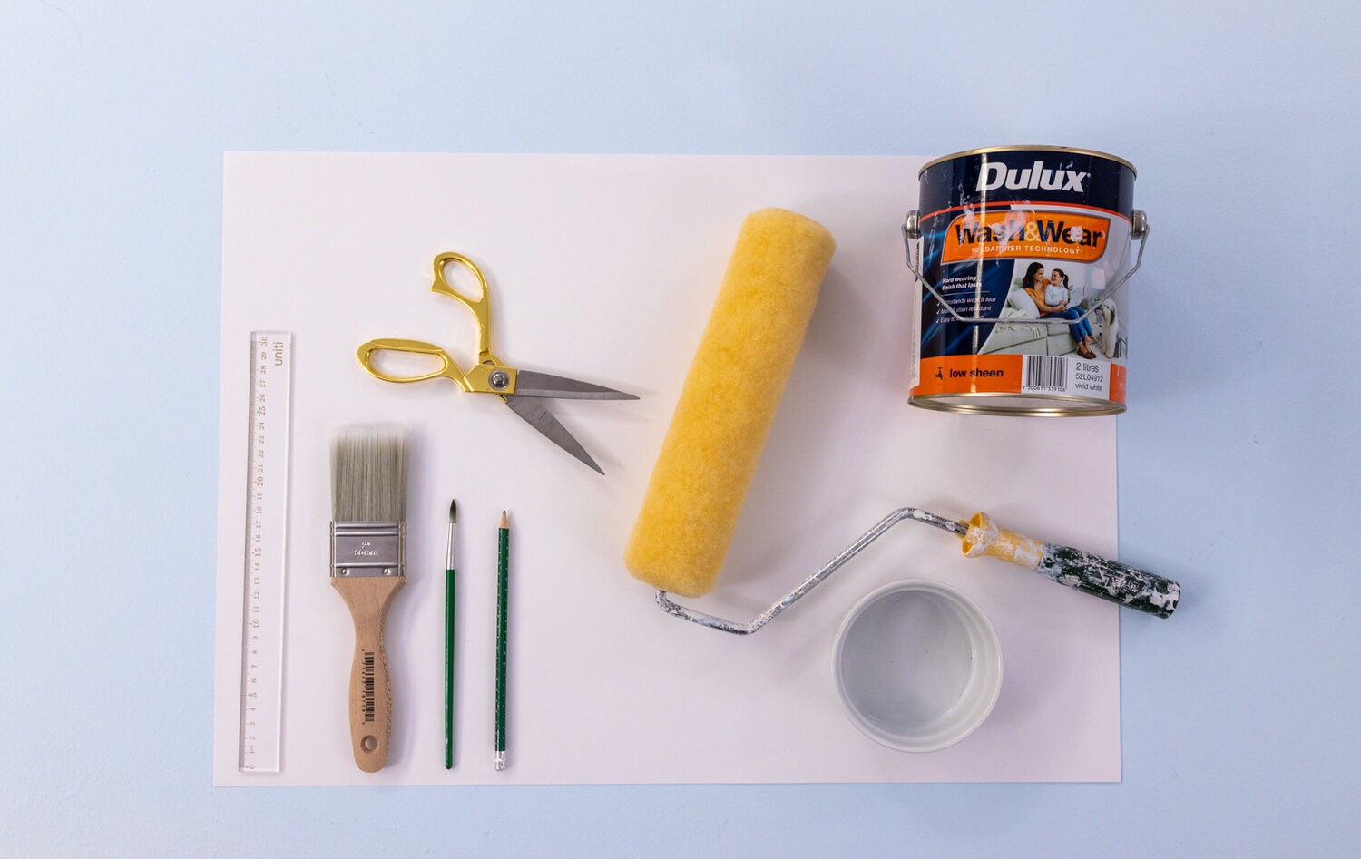 Supplies for the project lie on a table - a ruler, a large paintbrush, a small paintbrush, scissors, a roller, a ramekin, and paint.