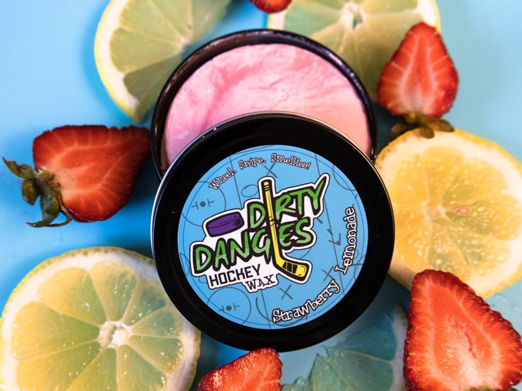 A tin of Dirty Dangles Hockey Stick Wax Strawberry Lemonade Scent sits open on a blue background surrounded by Strawberries and lemons
