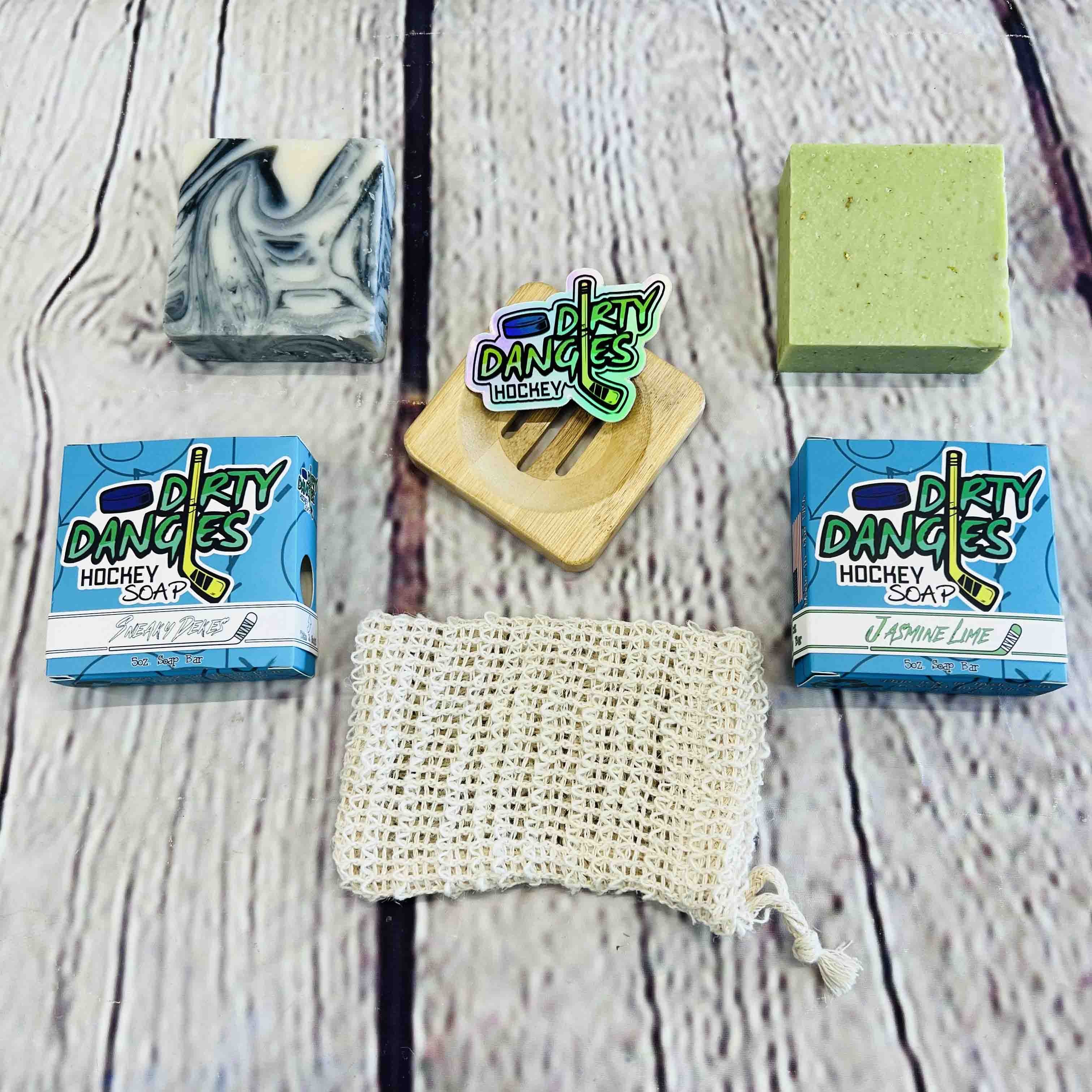 Dirty Dangles Hockey Soap Starter Kit Bundle. A black and white soap bar, sneaky dekes scent, a green soap bar jasmine lime scent, a tan soap bag, a bamboo soap dish and a dirty dangles logo sticker on a wood background.