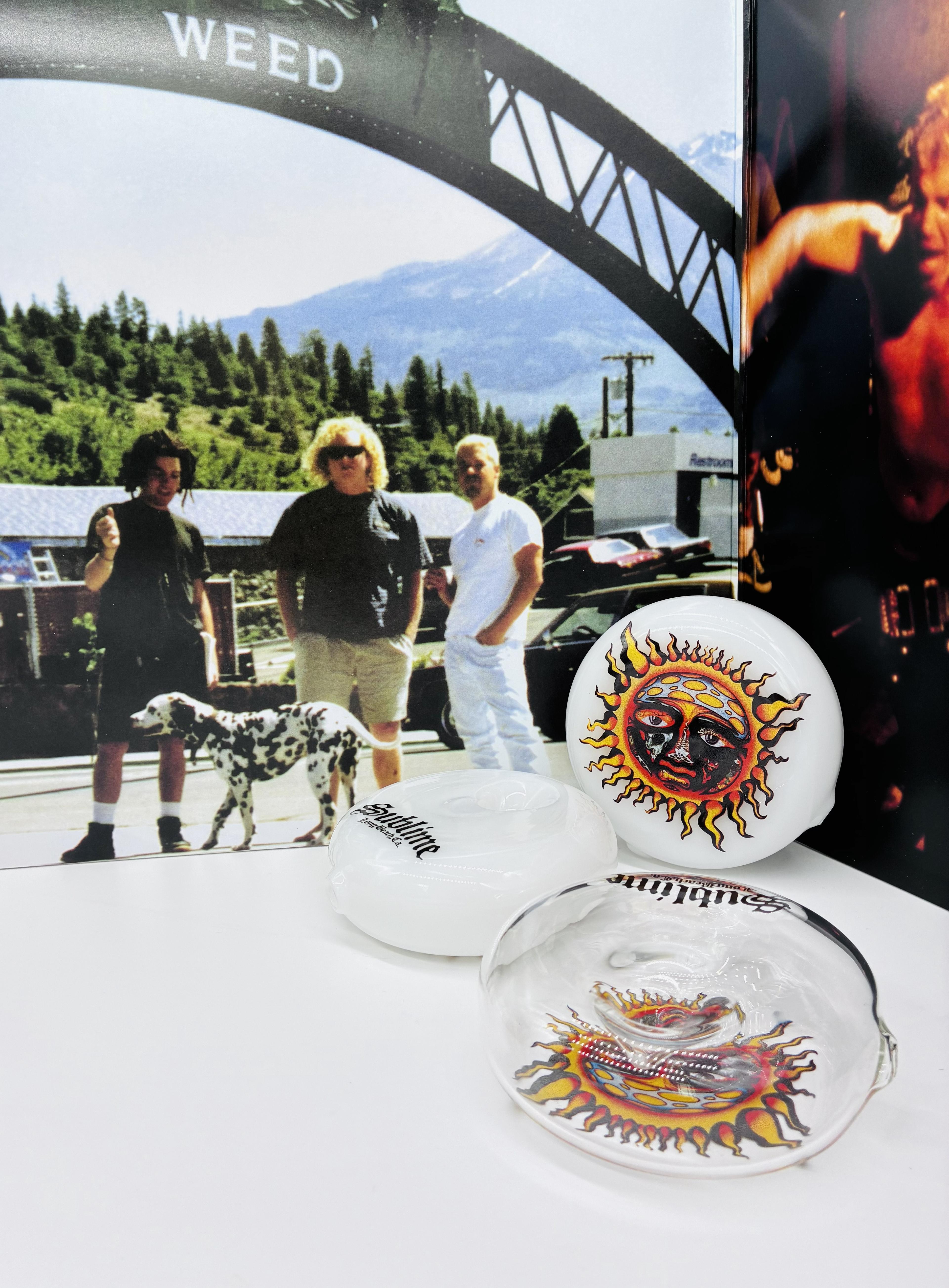 An image of three sublime handpipes, one clear and two opaque white ones with the iconic Sublime sun logo. the hand pipes are in front of propped up sublime vinyl albums and the album artwork, including an image of three men and a dalmation dog