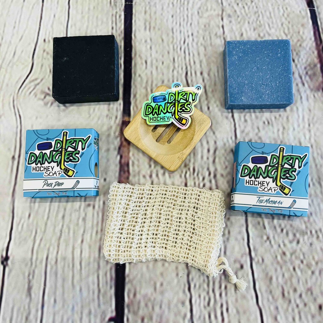 Dirty dangles hockey soap starter bundle on a wood background. A blue and black soap bar with their boxes, a bamboo soap dish, a soap bag and stickers.