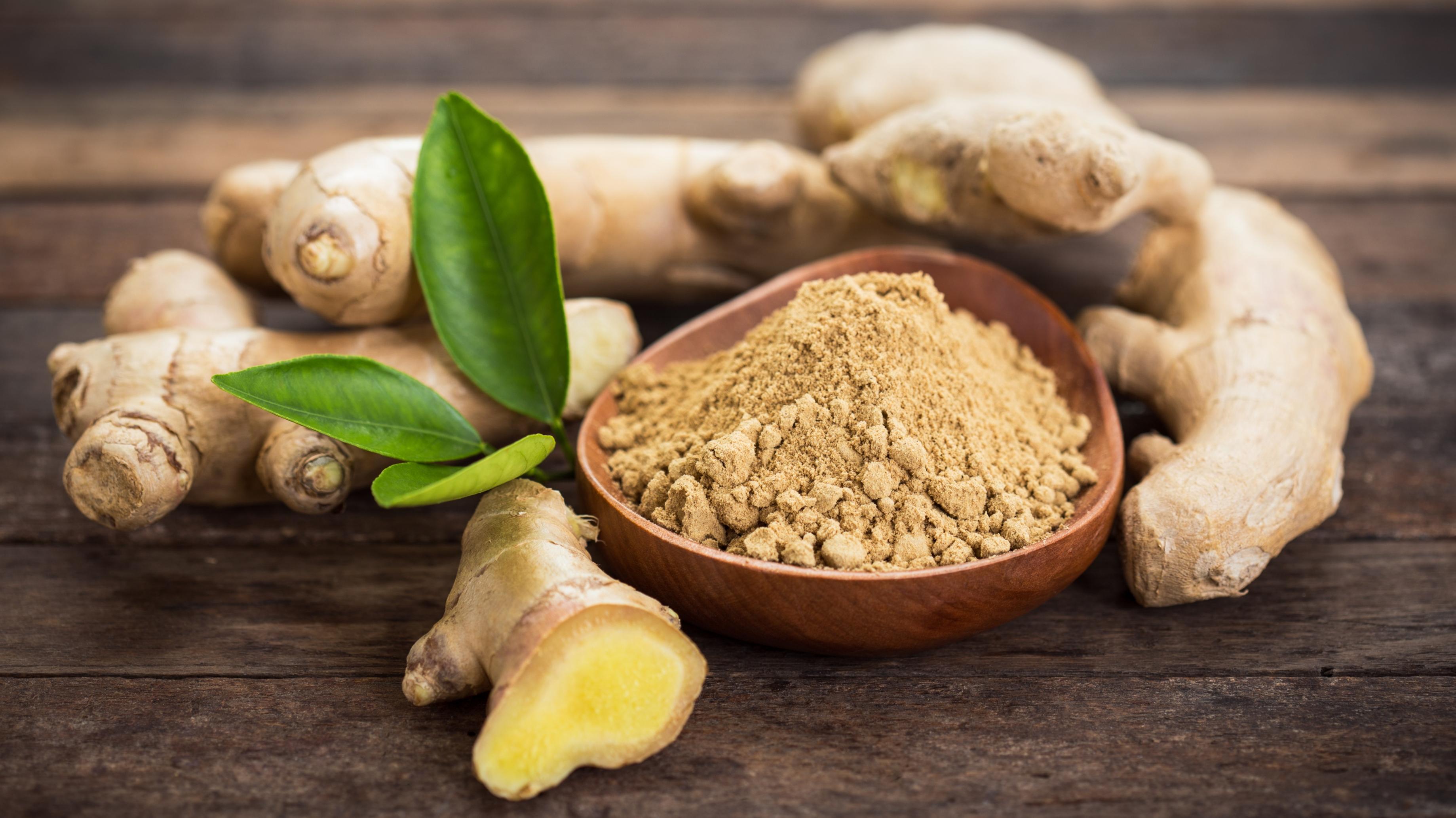 A bowl of powdered ginger with raw ginger surrounding the bowl and a green leaf on top of a brown wooden table.