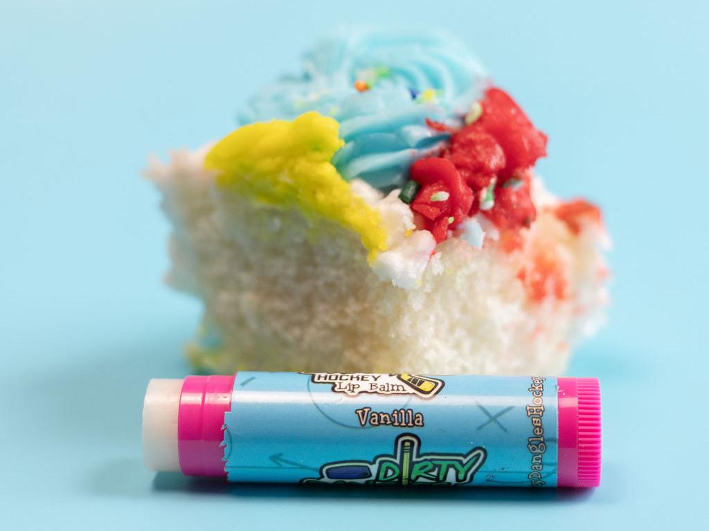 A pink stick of dirty dangles natural lip balm. Vanilla scent on a blue background with a piece of vanilla cake.