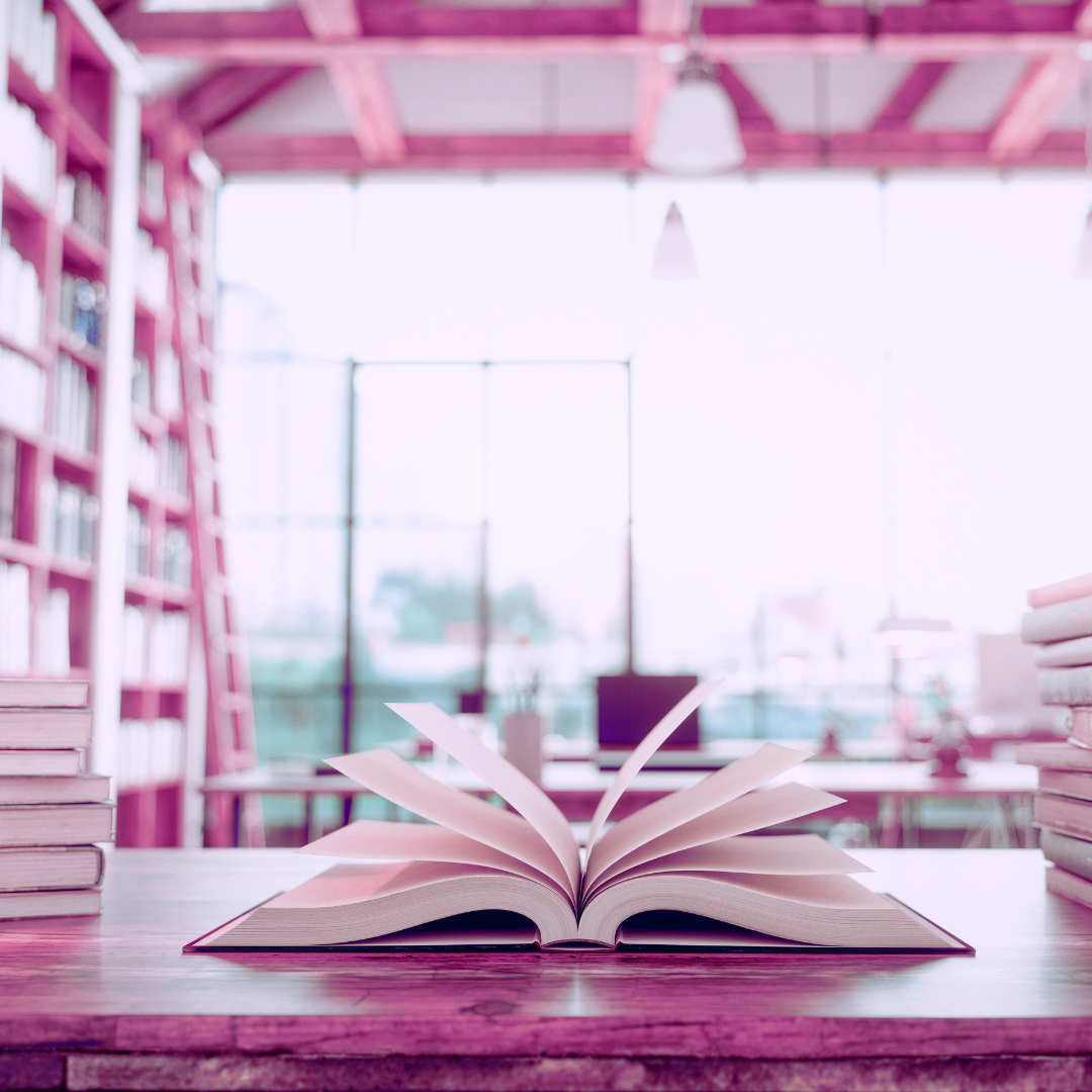 image of an open book on a desk in a library with stacks and shelves of books in a brightly lit room