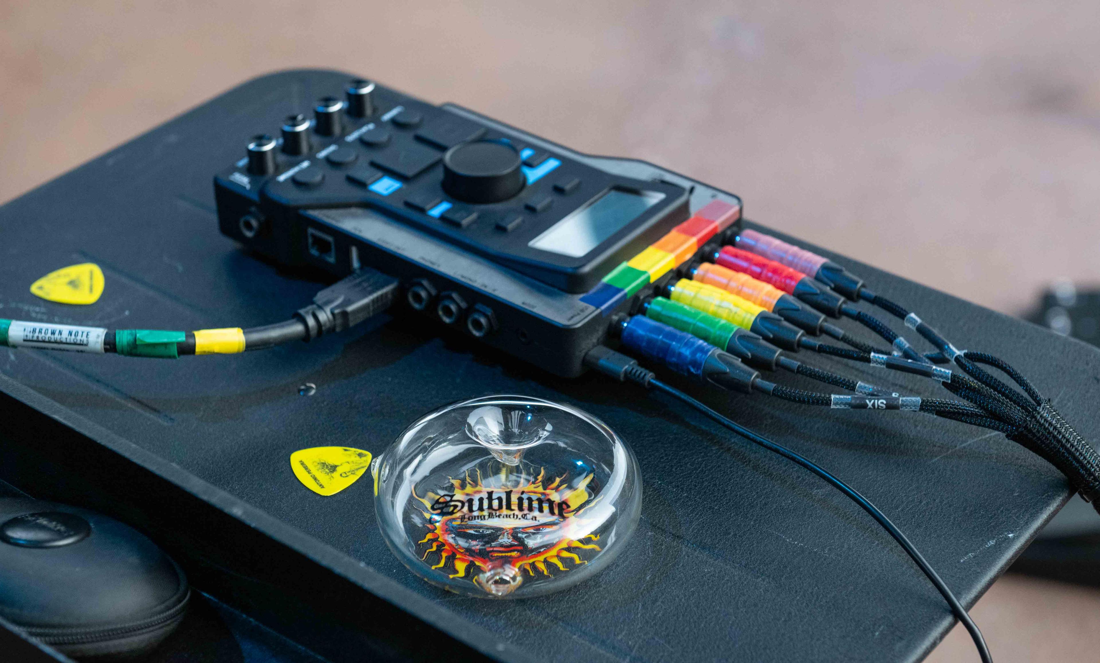 A clear and white Prism x Sublime donut-shaped glass handpipe sitting on a metal rack of music mixing gear backstage at a Sublime concert. The compact, circular pipe showcases the iconic Opie Ortiz Sublime Sun decal, highlighting the collection's portability, sleek design, and music culture roots.