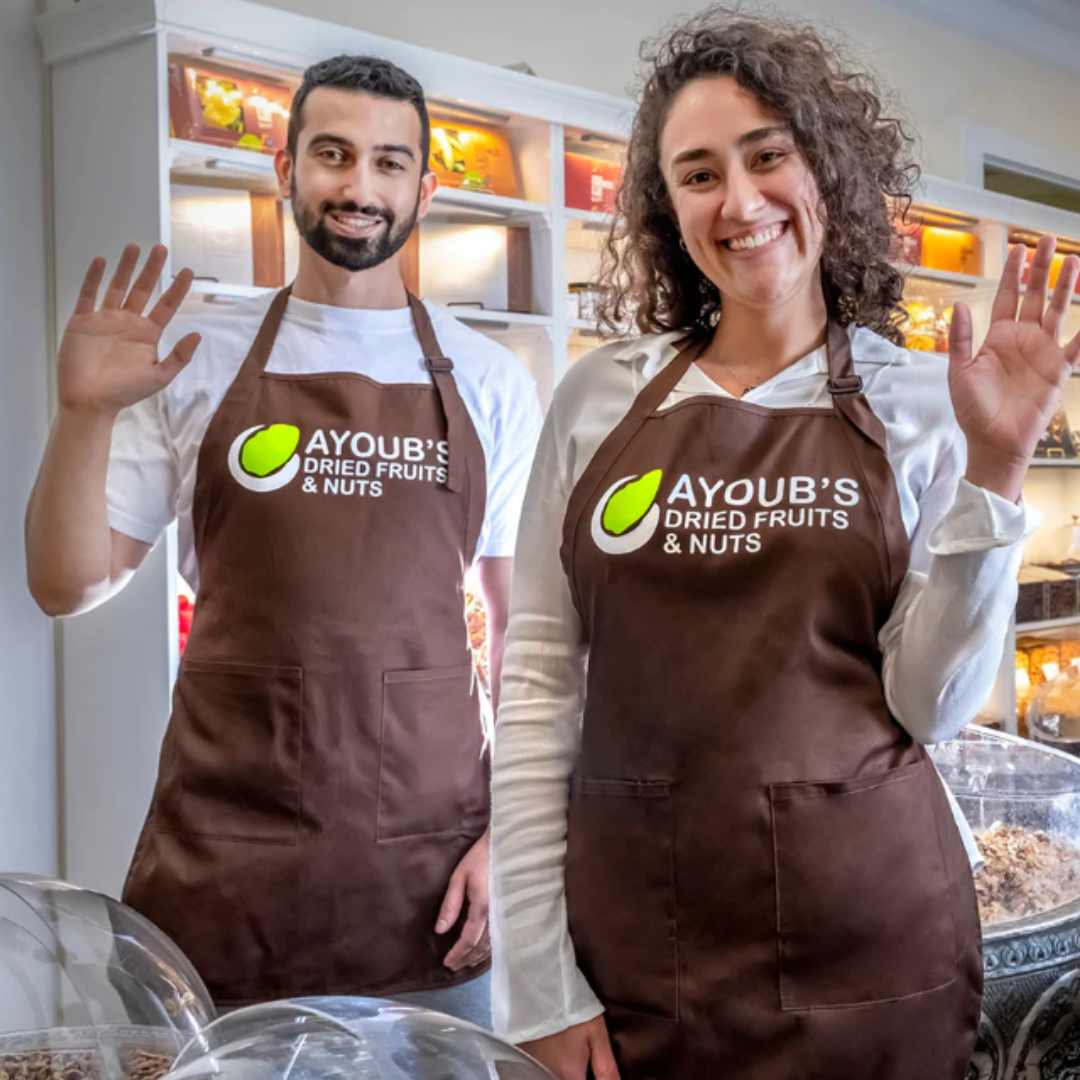 A smiling man and woman, both wearing brown aprons with the Ayoub's Dried Fruits & Nuts logo, wave at the camera while standing in front of shelves filled with dried fruits and nuts in a bright, welcoming store environment.