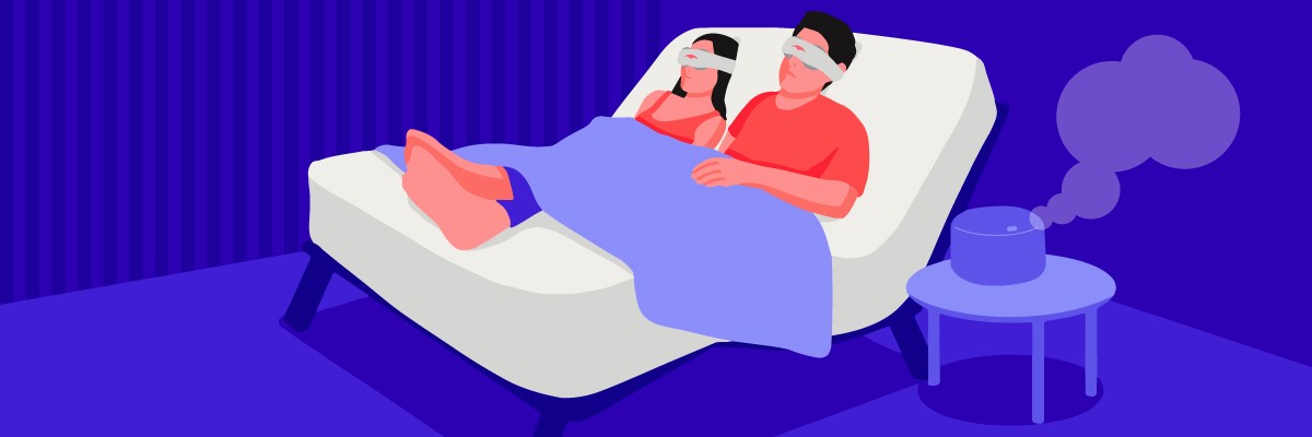 A man and a woman in sleep masks lying down on an adjustable bed while in the zero gravity sleep position. An aromatherapy diffuser sits on a night table beside the bed.