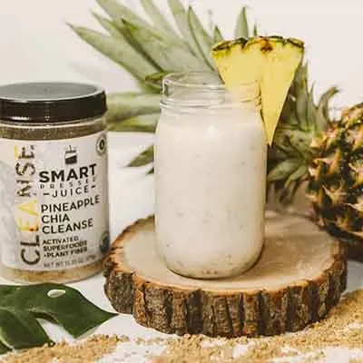A jar of the Pineapple Chia Cleanse set beside a wooden chopping board with a glass of white smoothie on top and a slice of pineapple wedge. There is a whole pineapple fruit behind and brown powder scattered in front. There is also a leaf displayed in front of the Pineapple Chia Cleanse jar.