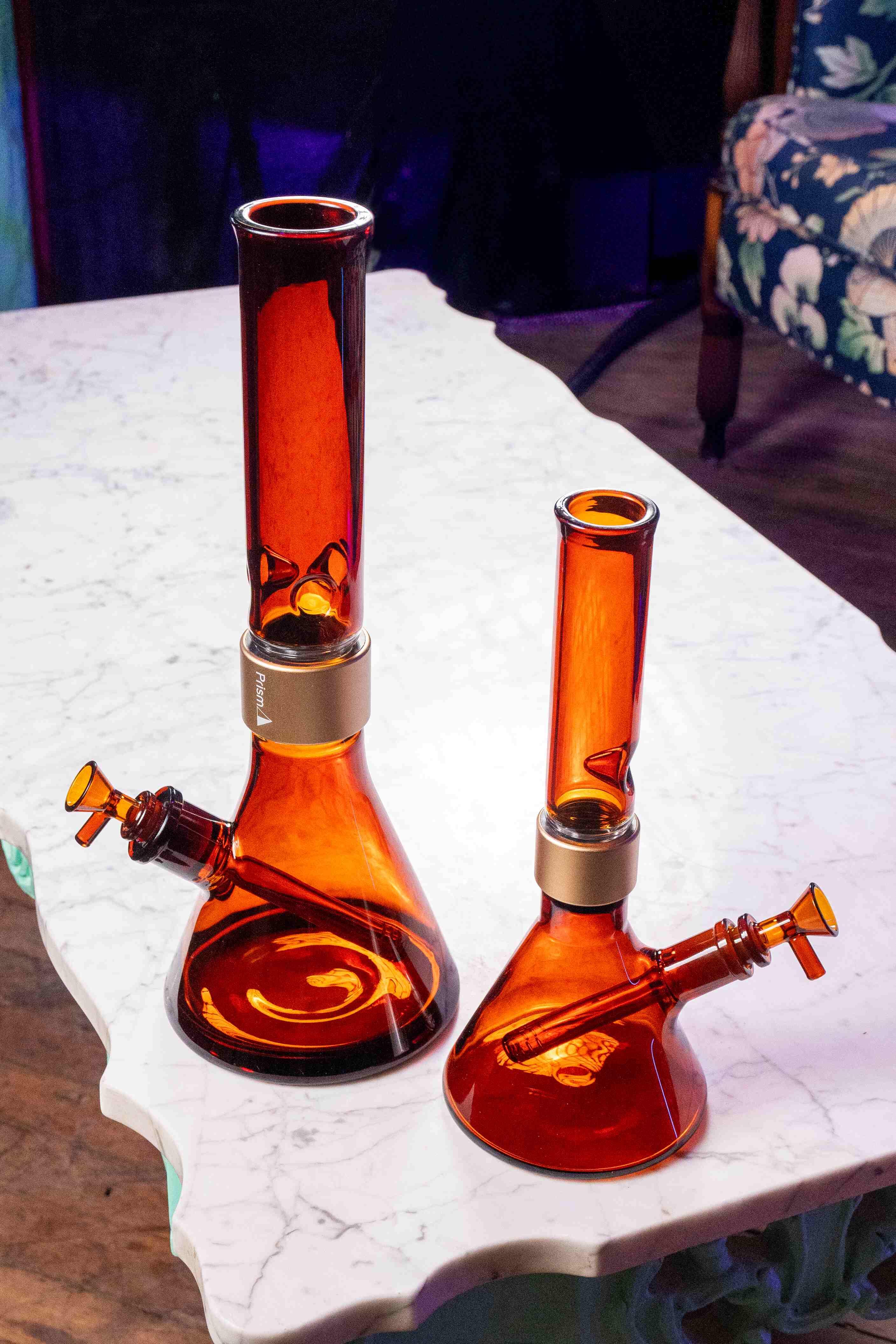 A side-by-side comparison of the Prism Amber Monochrome setups in Classic (left) and MINI (right) sizes. Both modular bongs feature amber beaker bases, matching amber bowls, downstems, and mouthpieces, all secured with signature gold halo connectors.