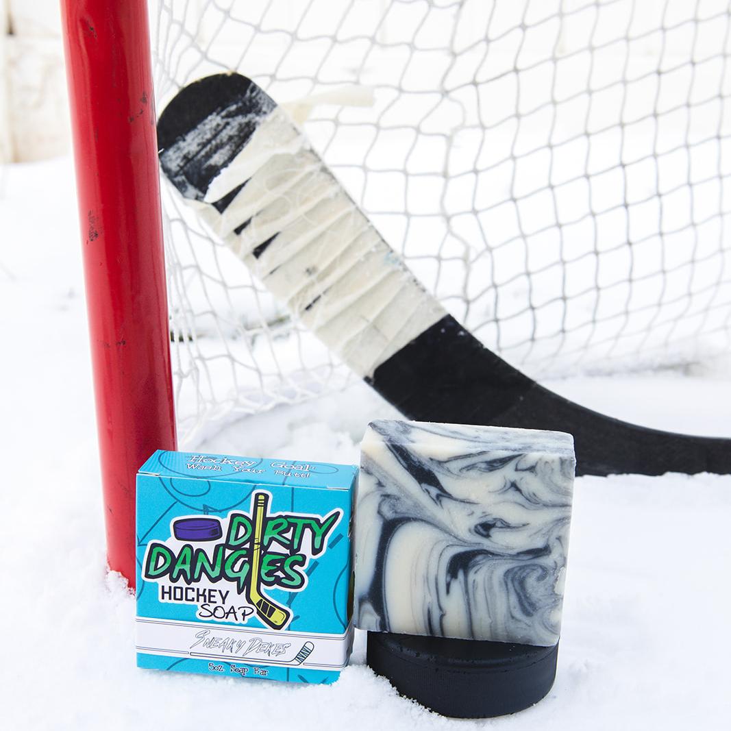 A black soap bar sits in the snow with a hockey goal, hockey puck and a hockey stick. Puck Drop