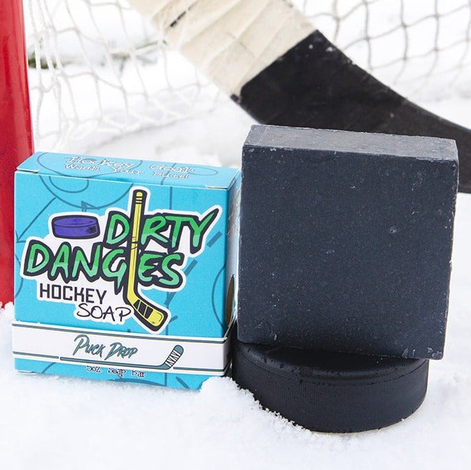 A black bar of dirty dangles hockey soap sits on a hockey puck in the snow with a hockey stick and a hockey goal. Puck Drop Scent