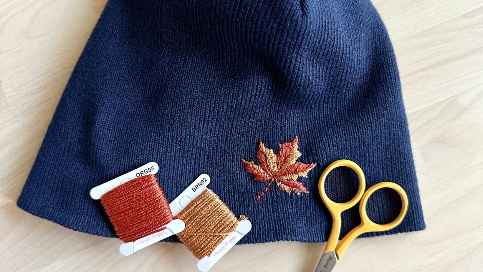 An autumn maple leaf embroidered on a navy beanie, with orange and brown floss cards and scissors.