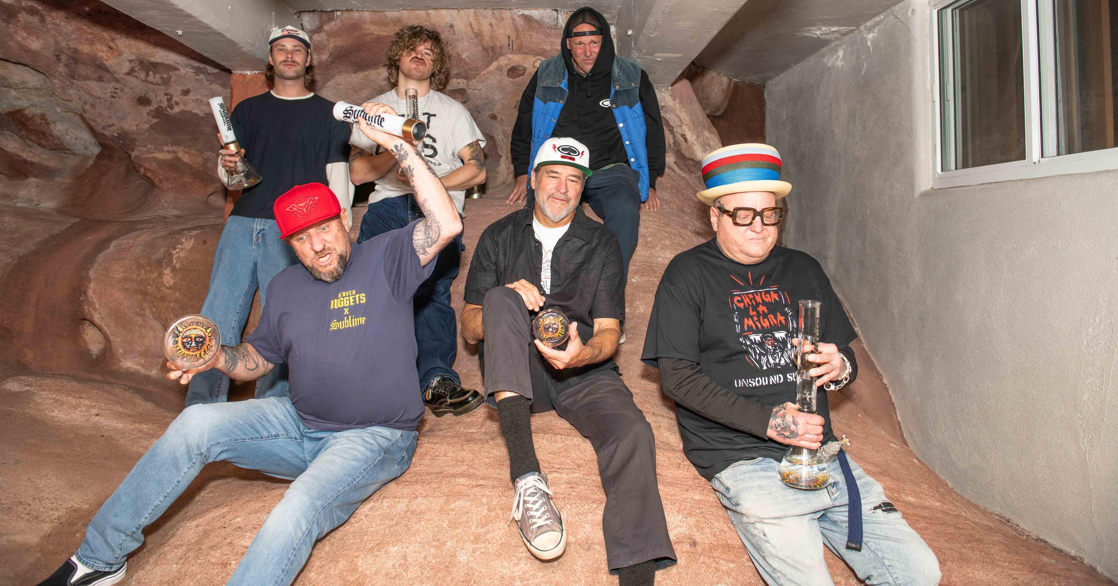 The full lineup of Sublime—including founding members Eric Wilson and Bud Gaugh, and frontman Jakob Nowell—posing at Red Rocks Amphitheatre. Each band member holds a piece from the official Sublime x Prism modular glass collection, celebrating the 2026 tour collaboration.