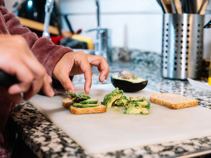 Hands preparing avocado toast, placing sliced avocado onto pieces of bread on a kitchen counter.