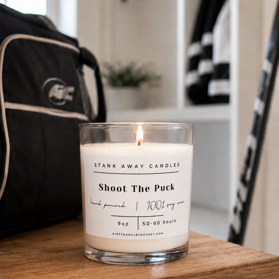 A white scented hockey candle on a wooden bench with hockey gear. Shoot the puck