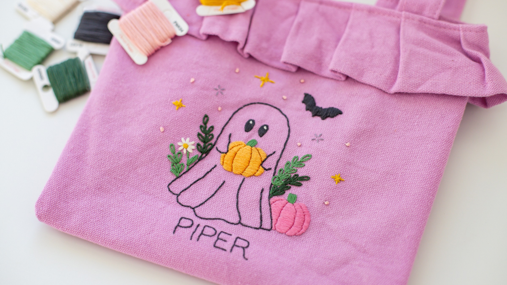A purple ruffle tote bag embroidered with a ghost holding a pumpkin and the name Piper.