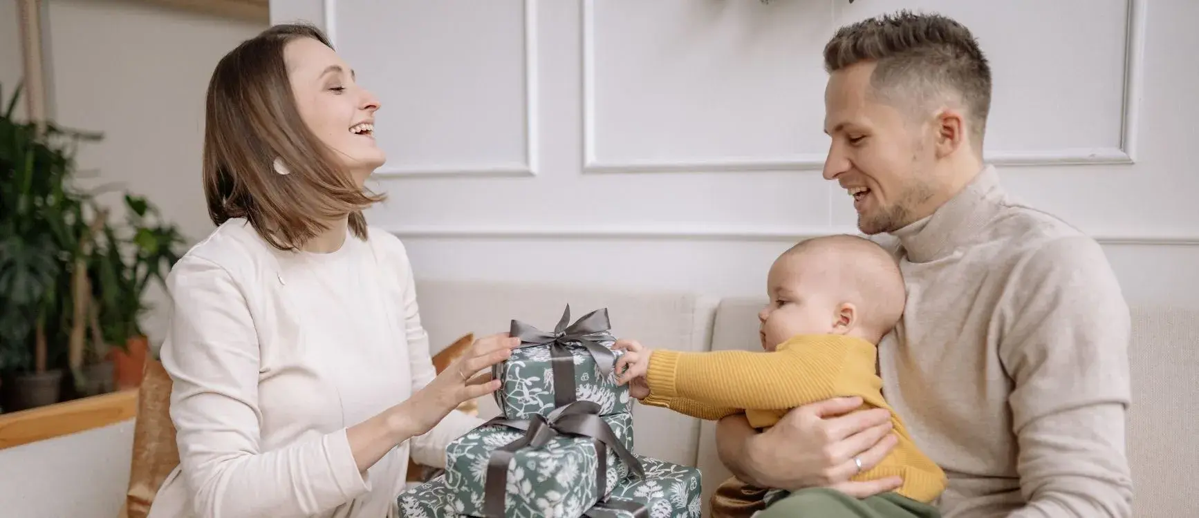 A woman and a man holding a baby sit on a couch as the baby reaches for wrapped gifts—showing how to build a baby registry without overspending, with smart, simple, and actually useful choices.