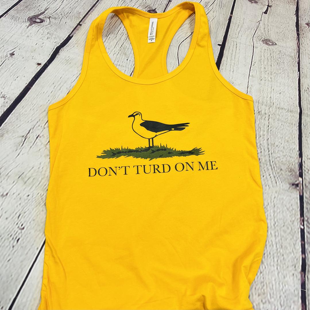 Picture of a gold and yellow women's tank top with a picture of a seagull standing in the grass. Text on the women's tank top says "Don't Turd On Me." Similar to the Gadsden "Dont Tread On Me" Flag